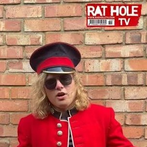 Still from Rat Hole TV: a band member in a red guardsman's jacket against a brick wall